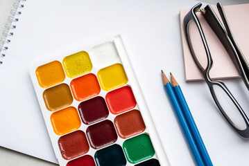 Colorful watercolor or acrylic paints, pencils and glasses on a white sheet of paper on a white background. Design studio table top view. 
