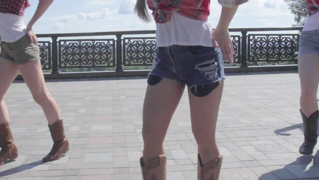 Several girls dancing country line dance on the concrete tile. Close up view to female hips and feet with brown cowboy country boots and jeans shorts. Traditional dance and style of America