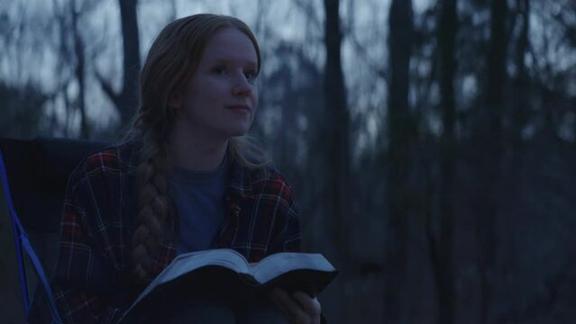 Young smiling woman sitting beside a camp fire and reading her Bible after sunset 