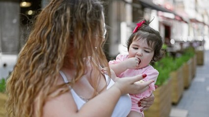 Smiling mother's confident expression as she's lovingly giving a salty snack to her happy daughter outdoors, casual family lifestyle in city street bathed in sunlight
