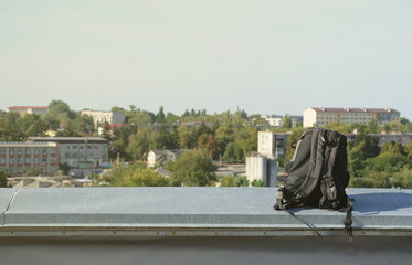 Black backpack lies on metal border of residental multistorey building rooftop in sunny weather outdoors