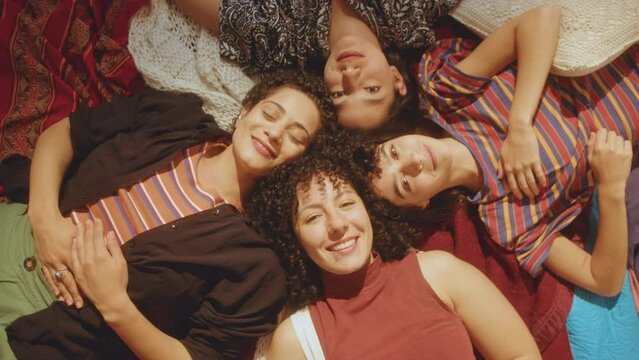 Company of young beautiful girls lying head to head on bed, looking at camera and happily smiling when hanging out at home. Directly above view, group portrait