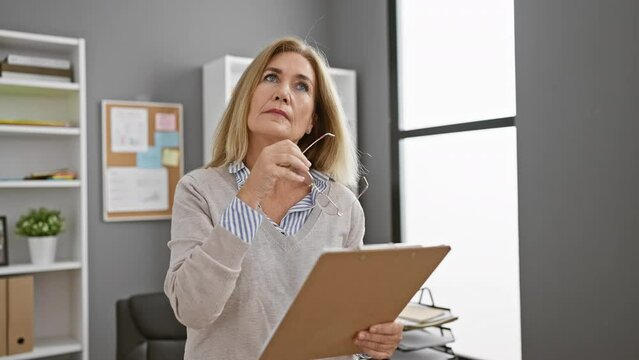 Middle-aged blonde woman in office contemplating with glasses and clipboard in a modern interior.