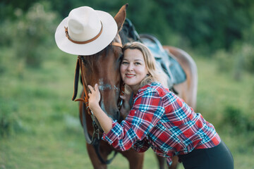 Happy blonde with horse in forest. Woman and a horse walking through the field during the day. Dressed in a plaid shirt and black leggings.