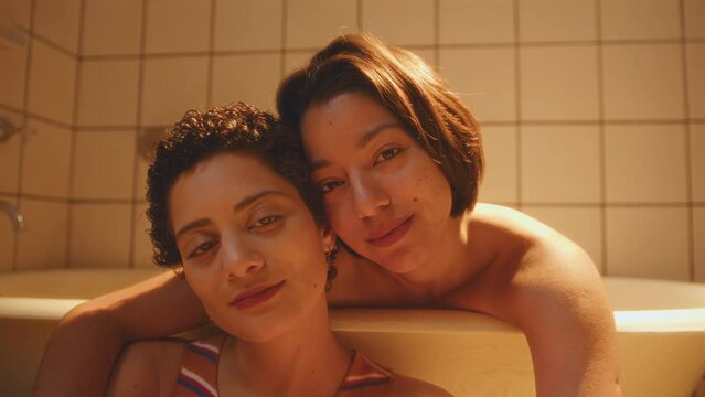 Young loving lesbian girlfriends embracing, touching each other with affection and posing on camera in the bathroom with warm light. Video portrait