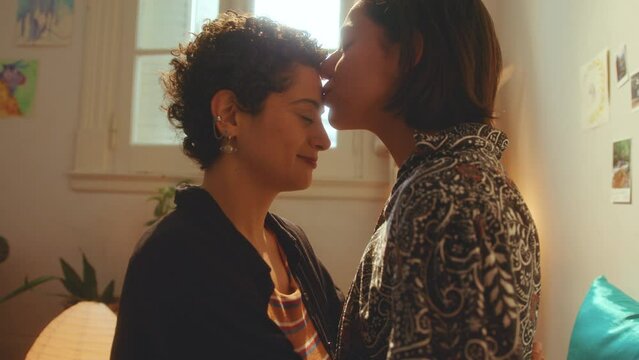 Young affectionate lesbian girls rubbing noses and kissing each other with love during the day at home. Zoom shot, side view