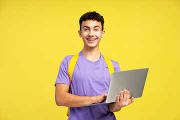 Portrait smiling teenage boy with backpack holding laptop computer isolated on yellow background....
