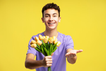 Smiling teenage boy holding bouquet of spring tulips looking at camera isolated on yellow...