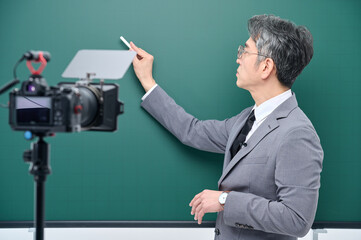 A middle-aged man is wearing a suit and giving a lecture in front of the camera pointing to the blackboard.