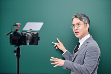 A middle-aged man is wearing a suit and giving a lecture in front of the camera pointing to the blackboard.