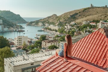 Woman sits on rooftop, enjoys town view and sea mountains. Peaceful rooftop relaxation. Below her,...