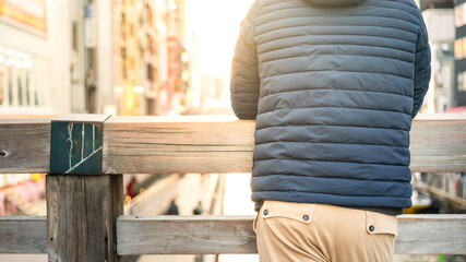A man wearing blue down jacket is standing and leaning on wooden rail with background of tourist city place. Photo applied with orange sunlight. - obrazy, fototapety, plakaty