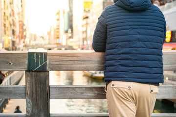 A man wearing blue down jacket is standing and leaning on wooden rail with background of tourist city place. Photo applied with orange sunlight. - obrazy, fototapety, plakaty