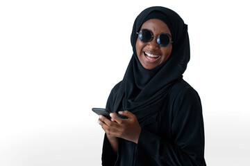 Happy African Muslim girl in black Hijab wears sun glasses smiling at camera, holding smart phone in hands on white background