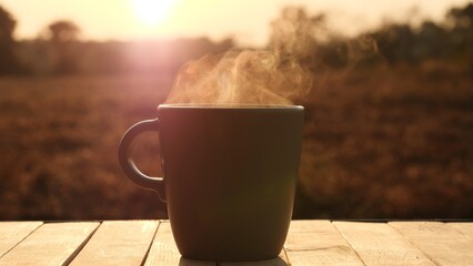 A close-up of beautiful vapor smoke rising from cup of coffee top wooden table light by morning sunlight against natural outdoor background, 4K. Concept of hot coffee, tea, breakfast, food, beverage