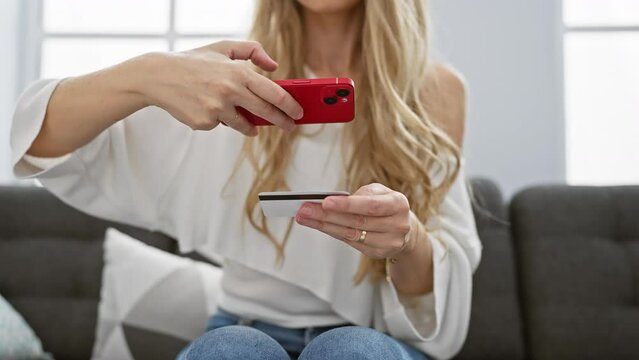 Blonde woman photographing credit card in modern living room interior.