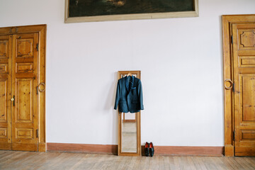 Groom suit hangs on a hanger on the floor mirror next to patent leather shoes in the large hall