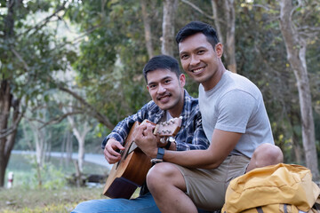 Asian LGBTQ couple enjoying nature, camping with tents in the forest area by the river, playing guitar.