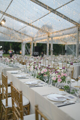 
Long table setting with beautiful flower arrangements for intimate wedding or party-outdoors family gathering on sunny day under transparent tent

