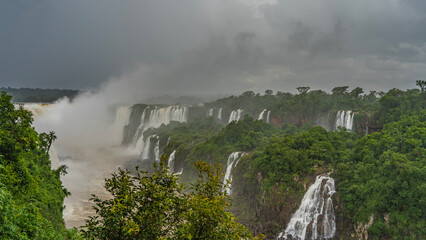 Many waterfalls collapse from ledges of rocks into the bed of a stormy river. Splashes, thick fog rise into the sky like a cloud. Lush tropical vegetation all around. Iguazu Falls. Brazil