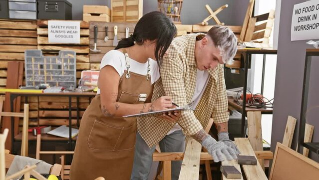 A man and woman team up in a woodworking workshop, as the man saws and the woman takes notes.