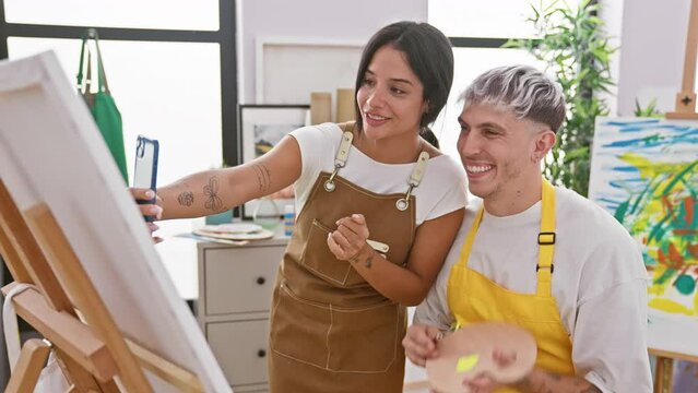 A man and woman in aprons smile while painting on canvas at a bright art studio.