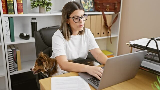 A focused hispanic woman works on a laptop in an office, with a pet dog by her side, depicting work-life integration.