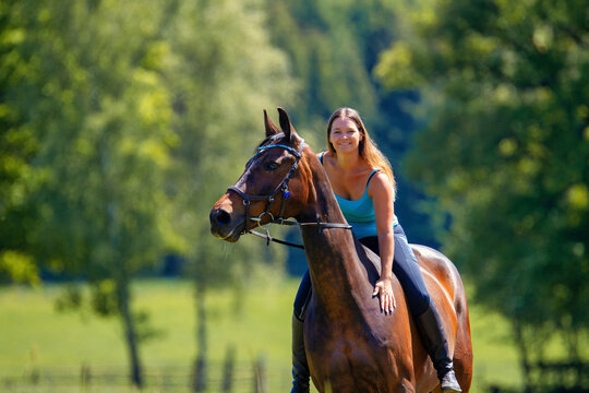 Young woman with brunette long hair rides bareback with her brown horse across a summer meadow, dressed in a blue tank top and riding pants with boots.