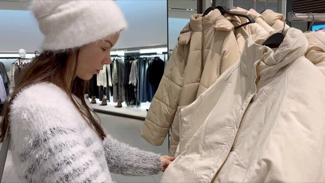 Young woman in white clothes choosing white down coat in a luxury boutique. Young adult woman looking for new clothes, shopping in a trendy brand apparel store. High quality 4k footage