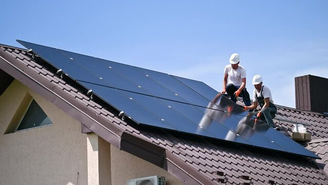 Workers building solar panel system on rooftop of house. Two men installers in helmets installing photovoltaic solar module outdoors. Alternative, green and renewable energy generation concept.