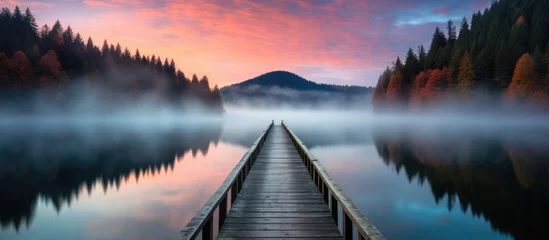 Keuken foto achterwand Reflectie A wooden pier extends into a foggy lake, reflecting trees, tranquil water, and a colorful sky.