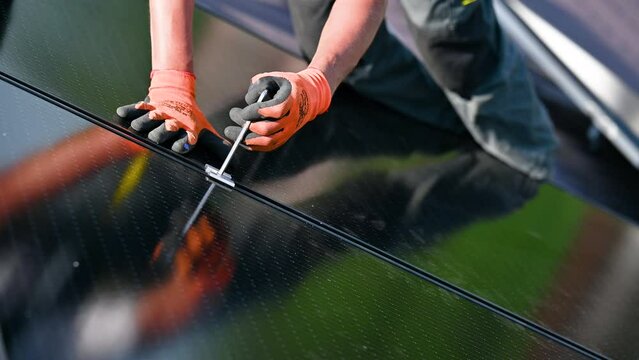 Worker building photovoltaic solar panel system on rooftop of house. Close up of man engineer in gloves installing solar module with help of hex key outdoors. Renewable energy generation concept.