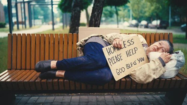 Elderly homeless woman lying on bench and holding please help cardboard sign