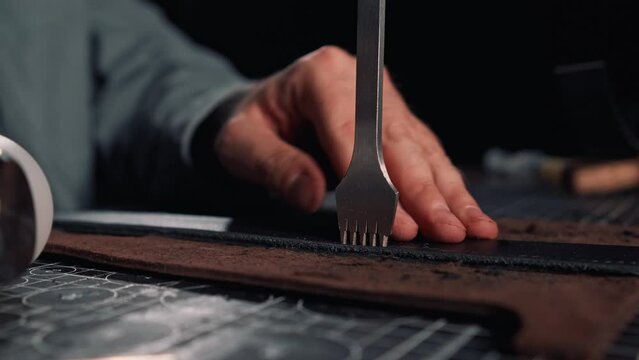 Close up of tanner hands making holes in leather purse using line piercer and hammer