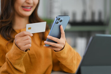 Satisfied young woman holding credit card using making online payments on mobile phone