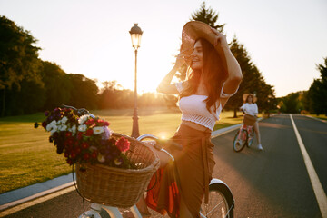 Female friends cycling together along local road at sunset - obrazy, fototapety, plakaty
