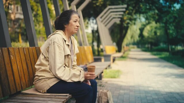 Old homeless woman holding paper cup while begging on bench, social inequality