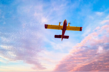 red and yellow amphibious firefighting aircraft flying in a sky of pastel blues and pinks
