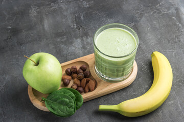 Green detox smoothie in a glass, blended vegetarian drink from spinach leaves, apple, banana fruit and nuts on dark background, healthy eating concept