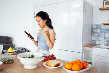Obraz na płótnie Canvas Young attractive Asian woman uses her phone and searches for recipe for dish on Internet. Charming female Korean cooks food in kitchen at home.