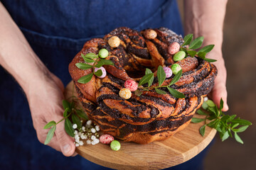 Man in a blue apron holding a freshly baked Easter cake with poppy seed filling and candies in a shape of eggs. Selective focus. Round yeast dough sweet bun. - obrazy, fototapety, plakaty