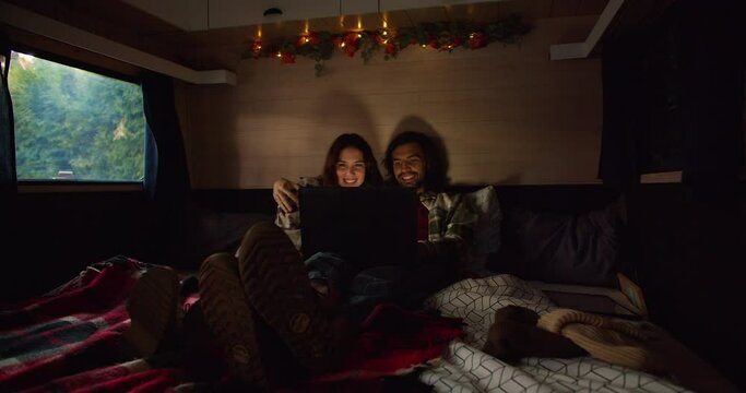 Shooting in an atmospheric trailer with dim lighting, a happy brunette couple in plaid shirts lie in their shoes on the trailer sofa and watch a movie through a laptop Near their camp outside the city