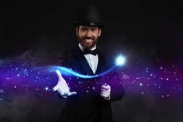 Smiling magician showing trick with wand on black background