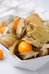 Ripe physalis fruits with calyxes in bowl on white marble table, closeup