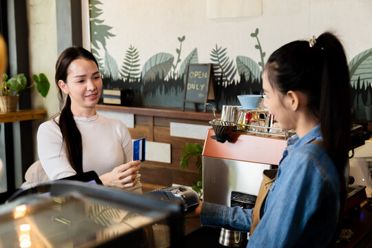 Asian waitress barista worker accepts credit card cashless payments from customer female in cafe restaurant, happy woman open bakery coffee shop, small business entrepreneur start-up lifestyle