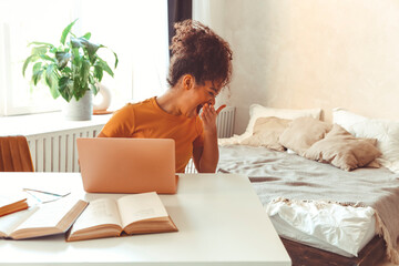 Tired flustrated African descent young girl sitting at desk in front of laptop while yawning cover...