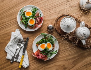 Delicious breakfast, brunch - toasted toast with arugula, boiled egg and prosciutto on a wooden table, top view