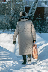 An elderly woman with a bag walks along a snow-covered sidewalk on a winter day