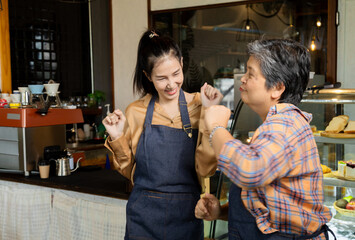 Young barista worker female and healthy senior waitress woman dancing in cafe counter bar small business restaurant, happy elderly 60s pensioner lifestyle working or business start-up entrepreneur