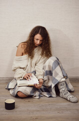 Beautiful girl is reading a book on the floor. Brunette with curly hair. Cozy home photo shoot.